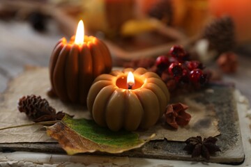 Burning candles in shape of pumpkins, old book and autumn decor on table, closeup
