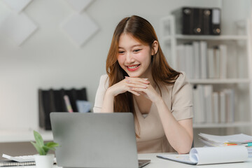 Focused Delight: Young Asian Businesswoman engrossed in her laptop at a modern office, her joyful...