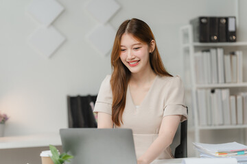 Focused and Productive: A young woman in a beige dress smiles brightly as she works on her laptop in a modern office setting. The image conveys a sense of focus, concentration, and professional succes