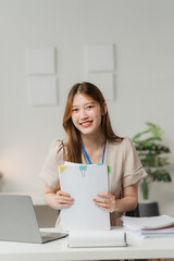 Confident and Organized: A young Asian businesswoman confidently holds a stack of paperwork in her modern office setting, exuding professionalism and efficiency.