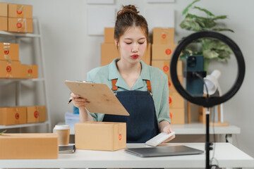 Livestreaming Logistics: Young Asian entrepreneur meticulously manages online orders, clipboard in...