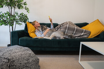 young woman in pajamas with her dog and with a blanket lying on the couch and using a smartphone