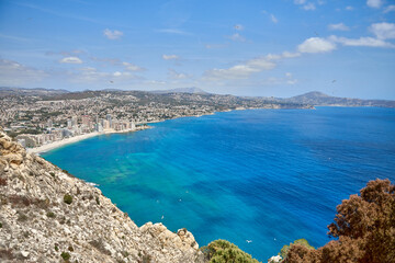 Breathtaking view from the cliff overlooking the stunning Calpe Bay in Spain with crystal-clear waters, sandy beaches, and majestic rock formations under a vibrant blue sky. Panoramic seascape of the