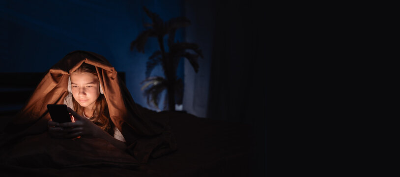 A teenage girl suffering from insomnia using a mobile phone for social media lies under a blanket on her bed in her bedroom - Powered by Adobe