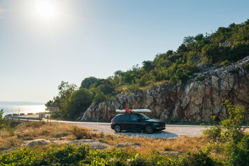 Black car with a paddleboard firmly strapped to its roof, parked on a cliffside road overlooking...