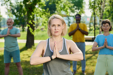 Naklejka premium Mature professional yoga instructor looking at camera while keeping her hands put together by chest during outdoor training