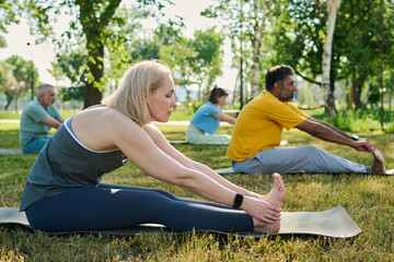 Mature blond woman in activewear sitting on mat and bending forwards while teaching elderly people...