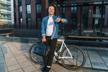 Obraz premium A smiling man stands with his bicycle, checking his watch, dressed casually, holding a briefcase.