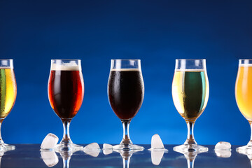 Glasses with different types of beer and ice cubes on mirror surface against blue background