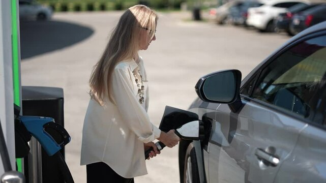 Happy mature businesswoman unplug charger plug from charging station attach to electric car before driving around city center. Eco friendly rechargeable car powered by sustainable and clean energy