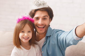 Portrait of pretty little princess and her happy father wearing crown, playing together at home, taking selfie, copy space