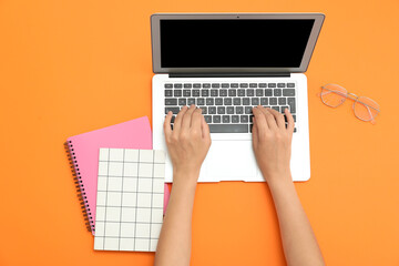 Fototapeta premium Female hands with laptop, notebooks and eyeglasses on orange background