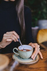 Close-up picture of pretty girl's hands in a black sweater gently stirring her tea in a light green cup, surrounded by a warm cafe environment filled with plants.