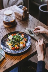 A beautifully arranged salad on a dark plate features vibrant cherry tomatoes and greens. A person sits at the table, ready to enjoy their meal.