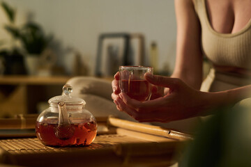 Hands of tranquil woman in activewear holding glass of hot herbal tea over bamboo tray with teapot...