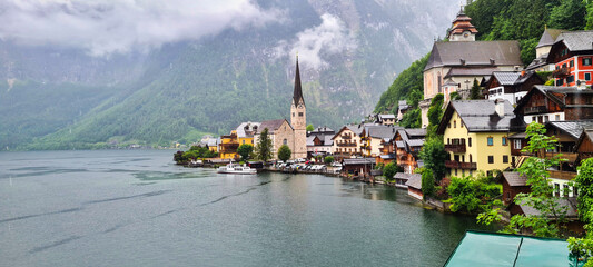 A picturesque view of Hallstatt, Austria, nestled by the tranquil lake and surrounded by towering misty mountains. The charming village is adorned with colorful houses and a prominent church, making i