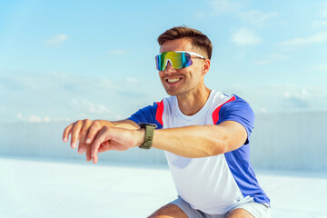 A man smiles while performing a squat outdoors, wearing sunglasses and sportswear, enjoying the...