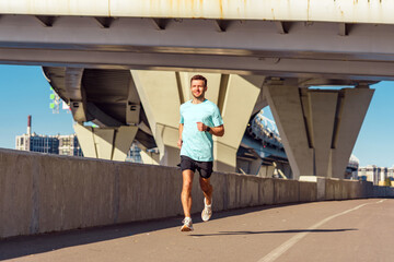 A man runs confidently along a path beneath an urban bridge, enjoying a sunny day and staying active.