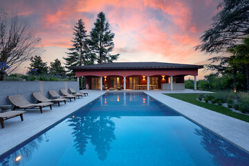 House with a nice pool in front, sunset photography. Lots of deck chairs on the left. Fire red sky, all in the middle of nature.