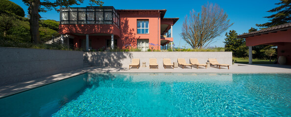 Front view of a modern villa with a huge swimming pool. On the opposite side there are six deck chairs lined up for sunbathing and the sky is blue.