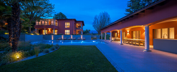 Front view of a modern villa with outbuilding. There is a large table for dining outside and a large swimming pool for relaxing. No one inside, night scene