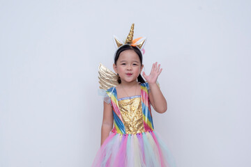 A joyful little asian girl dressed in a colorful unicorn costume with a golden horn and wings, smiling playfully and waving hi. Isolated on a white background.
