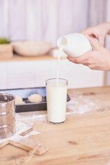 Pouring fresh milk into a glass on a wooden kitchen table with baking ingredients. Child portrait. People baking