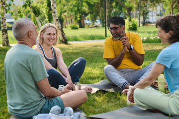 Naklejka premium Group of cheerful mature intercultural men and woman sitting on mats in front of female yoga instructor and having fun during chat