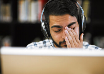 Man, university student and tired on computer with headphones in library for internet, online error and research. Male person, exhausted and burnout or headache with assignment deadline and study