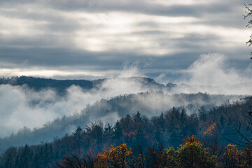 Fototapeta premium Serene view of foggy hills in the early morning, with a forest draped in a dense layer of mist. The fog rolls through the trees, creating an ethereal and calming atmosphere under a moody, overcast sky