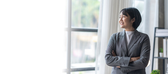 Fototapeta premium Young businesswoman smiling and looking out the window with arms crossed in her office