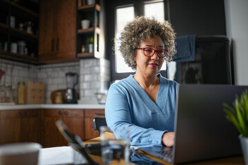 A mature woman with curly gray hair and glasses works on a laptop in a cozy home kitchen. The...