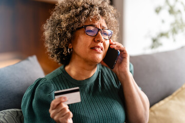 A shocked woman with curly hair and glasses is holding a credit card while talking on her phone,...