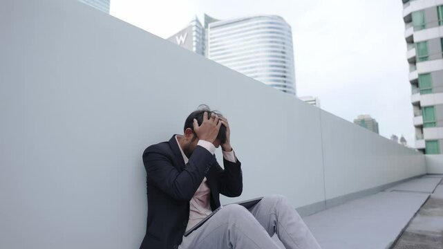 A businessman in a suit sits on the ground outside a modern building, holding his head in frustration. The scene reflects stress, anxiety, or challenges in a corporate environment.
