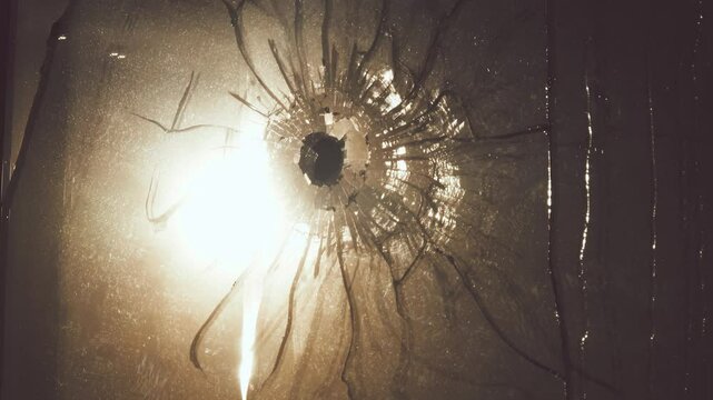 Bullet hole is in double pane of glass. Web of cracks, old web of spider debris is stirring in wind. Sweat streaks. Reflection of harsh sun in window. Concept of troubled world, war enters every home