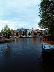 peaceful Amsterdam canal with traditional Dutch houses, parked bicycles, and boats on calm water. Arched bridges and green trees frame the scene, warm sunset light reflecting on historic buildings