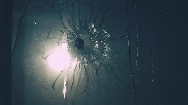 Bullet hole is in double pane of glass. Web of cracks, old web of spider debris is stirring in wind. Sweat streaks. Reflection of harsh sun in window. Concept of troubled world, war enters every home