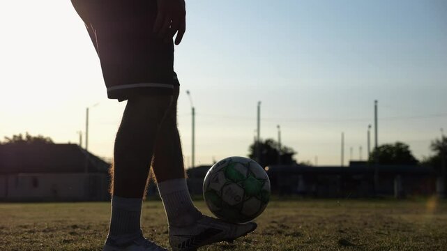 Legs of young man kicking ball at green field. Male feet of professional footballer juggling soccer ball on stadium at sunset. Sportsman practicing tricks outdoor. Concept of a freestyle football