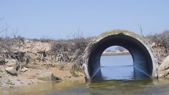 A concrete pipe (culvert) under a country road. The flow of water. The outskirts of the village are visible through the opening of the pipe