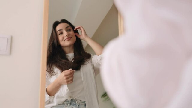 A young woman looks into a mirror, smiling, exuding positivity and high self-esteem in a bright, comfortable room.