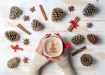 Woman's hands holding mug of coffee with cinnamon, star anise and pine cones over white wooden table. Christmas or New Year holiday background. Flat-lay.