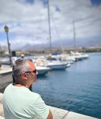 Profile of a Caucasian adult man sitting, looking at the port. Man looking at the sea. Boy sitting alone looking at the small port. Observing a small port. Tourist in Tenerife sitting admiring