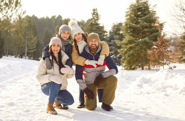 Happy family with kids standing in winter park together hugging and looking at camera. Young parents with two children walking outdoors in snowy forest. Family leisure and winter holidays concept.