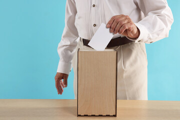 Referendum. Man putting his vote into ballot box at wooden table against light blue background,...
