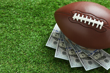 American football ball and dollar banknotes on green grass outdoors, space for text