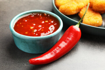 Hot chili sauce in bowl and pepper on grey textured table, closeup