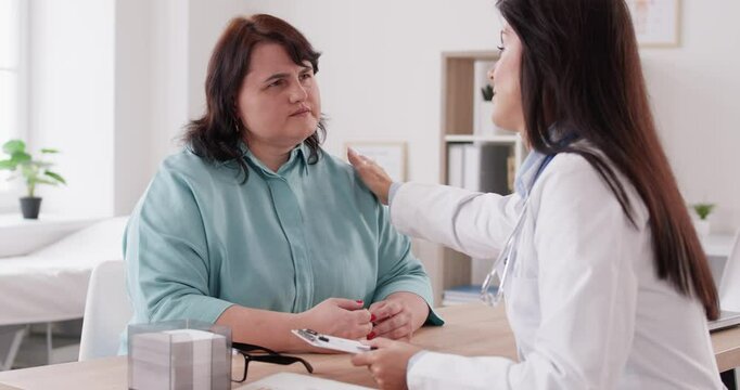 Middle-aged Caucasian woman at doctor appointment. Female overweight patient telling about her health issues. Nutritionist taking notes and encouraging patient during consultation in medical office.