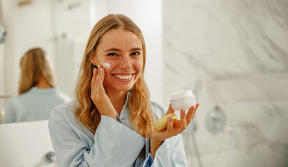 In her stylish bathroom, a joyful woman applies skincare products and radiates positivity...