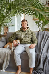young man smiling looking at the camera with his pet listening to music with headphones on in the living room of his house, vertical, palm tree, sofa, peace, relax, tranquility, best friend,