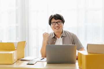 Middle aged Asian entrepreneurs using laptop computer. Mature Asian man sitting packing canvas in box to customer and check order with computer. Small business owner. SME e-commerce concept.